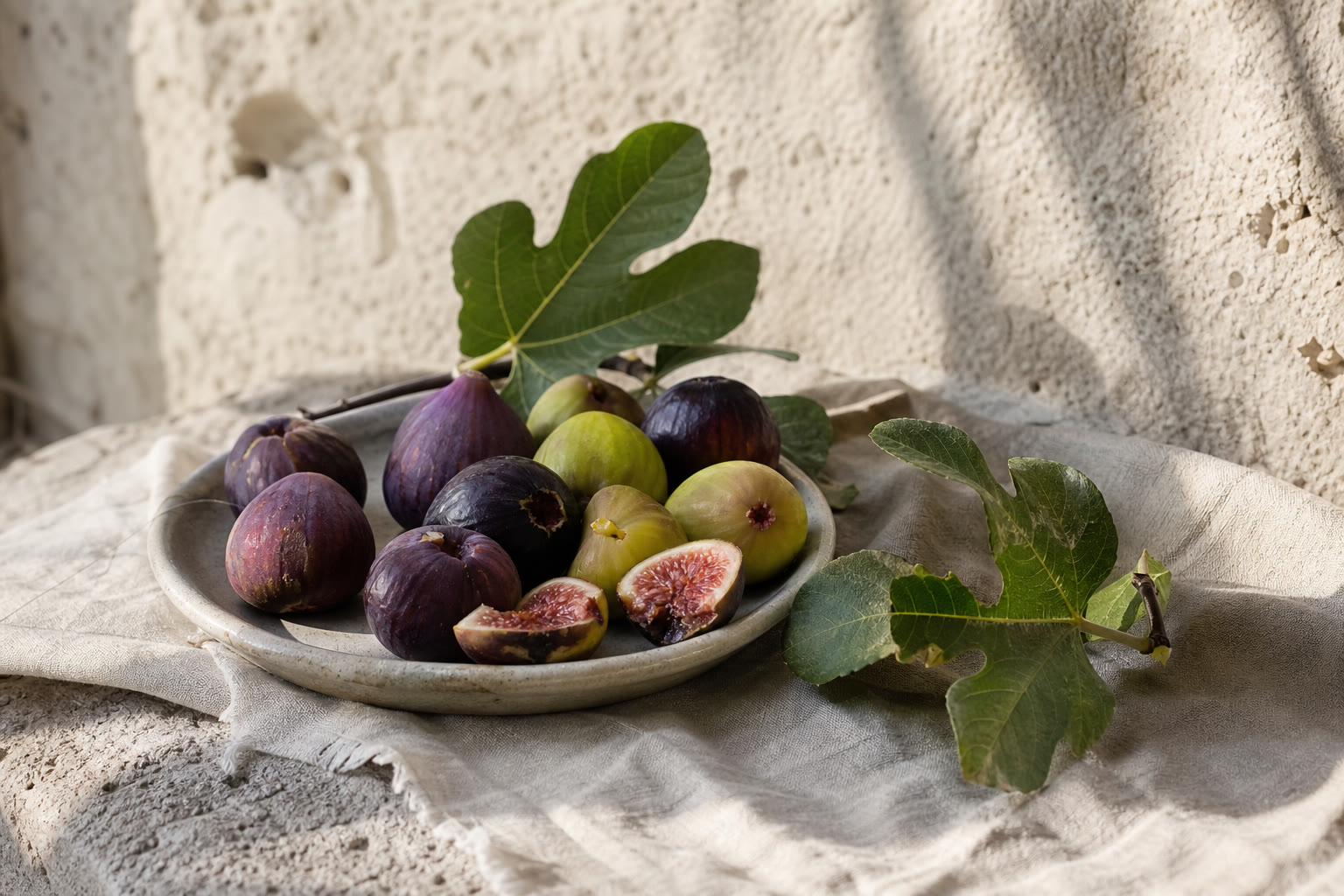 Fichi italiani maturi appena raccolti su pietra e lino naturale, due aperti a metà, nella luce morbida di fine estate