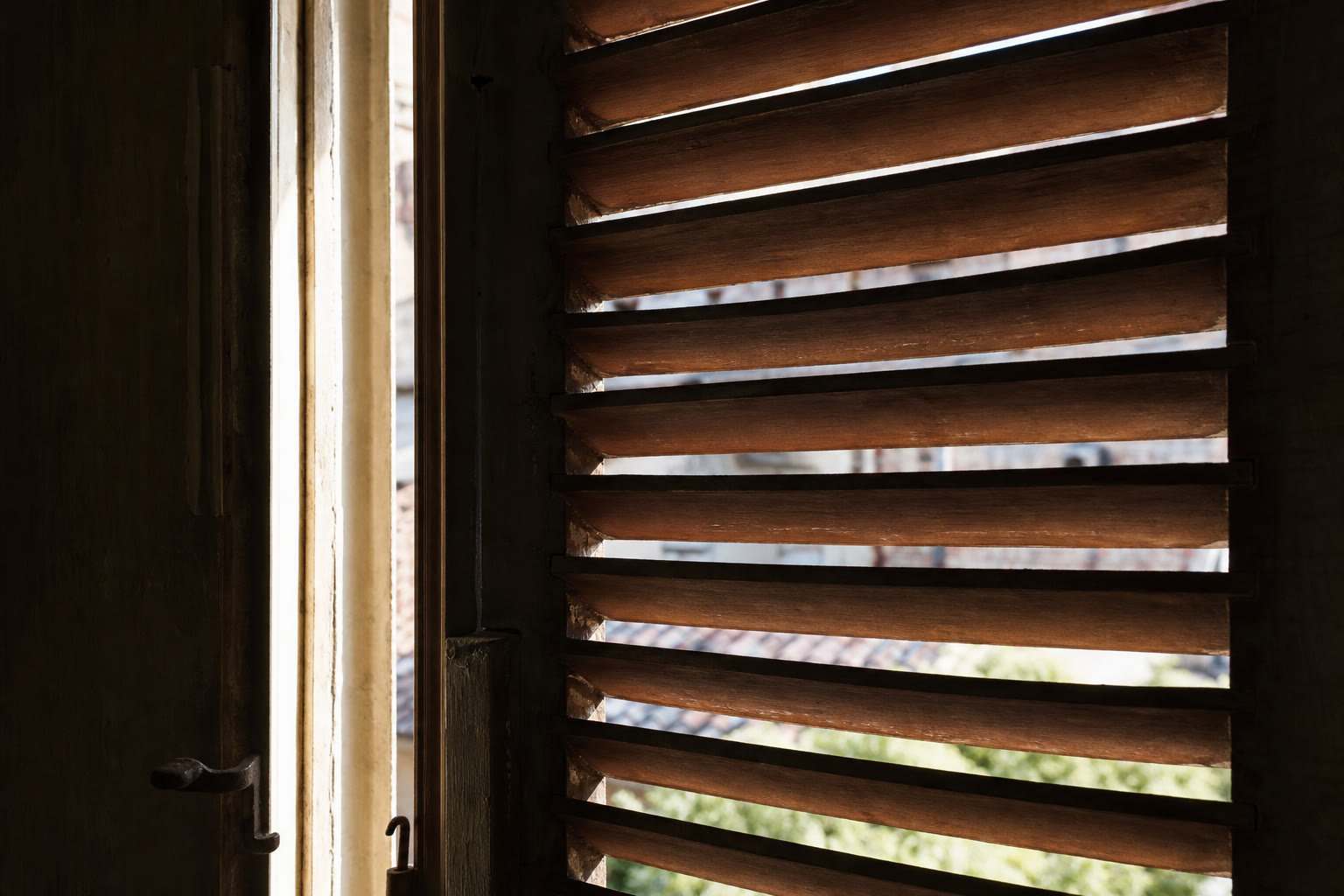 Dettaglio ravvicinato di una persiana tradizionale italiana in legno semiaccostata, con le lamelle che filtrano la luce intensa dall'interno